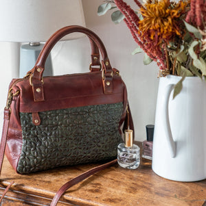 Quilted fern handbag on a dresser next to a vase of flowers and some perfume and nail polish, Rosalie Quilted Crossbody Bag.