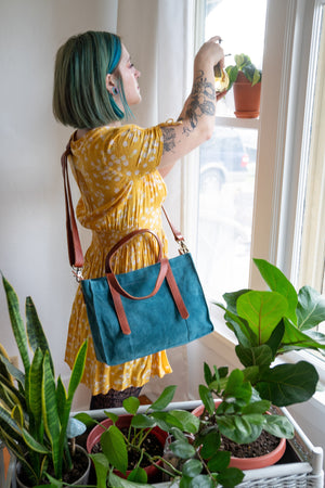 Square teal suede tote being worn as a crossbody bag on a woman watering plants, Aurora Suede Tote.