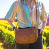 Brown woven cross-body bag on a woman standing in a field of flowers, Sawyer Woven Shoulder Bag.