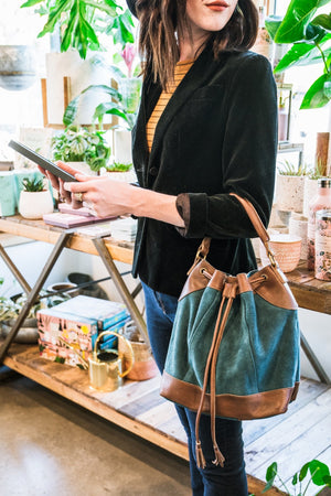 Woman standing in a plant shop wearing a teal suede crossbody bag, Rowan Suede Crossbody Bag.